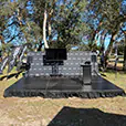 Outdoor event stage setup with large TV screen, speakers, microphone stand, and branded backdrop surrounded by trees in a park setting