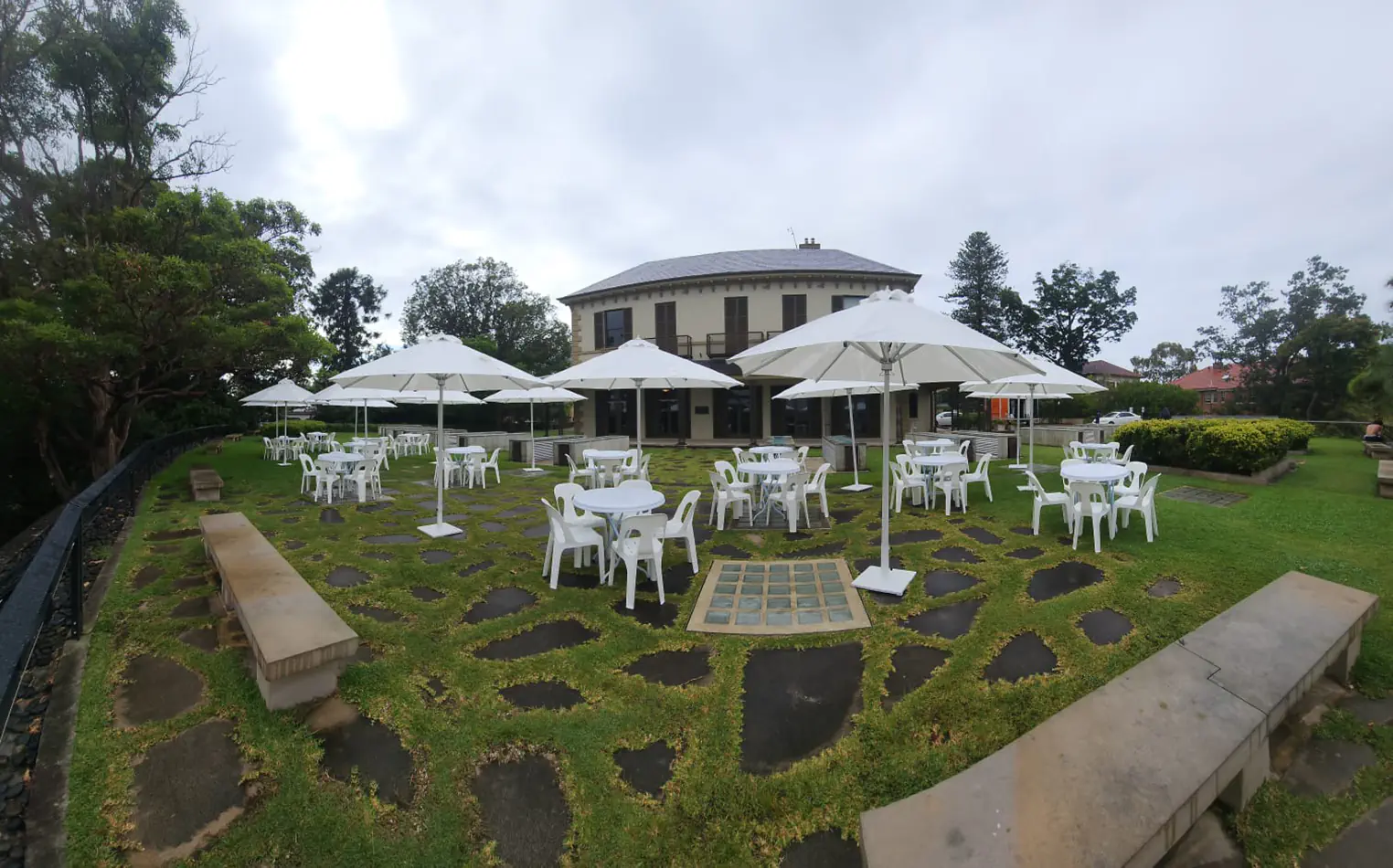 Outdoor event setup with round white tables, plastic chairs and large white umbrellas arranged on a garden lawn near a building