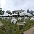 Outdoor event setup with round white tables, plastic chairs and large white umbrellas arranged on a garden lawn near a building