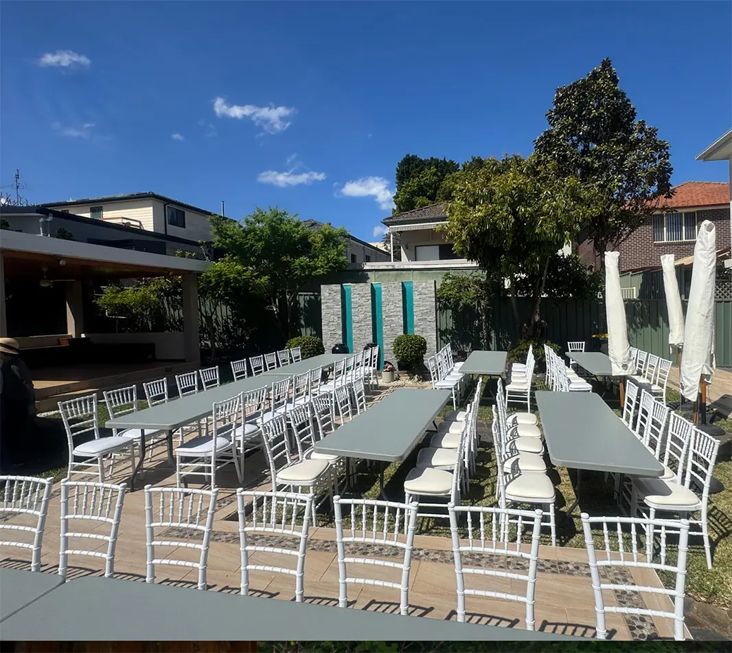 Outdoor event setup with long grey trestle tables and white Chiavari chairs arranged in a private backyard venue