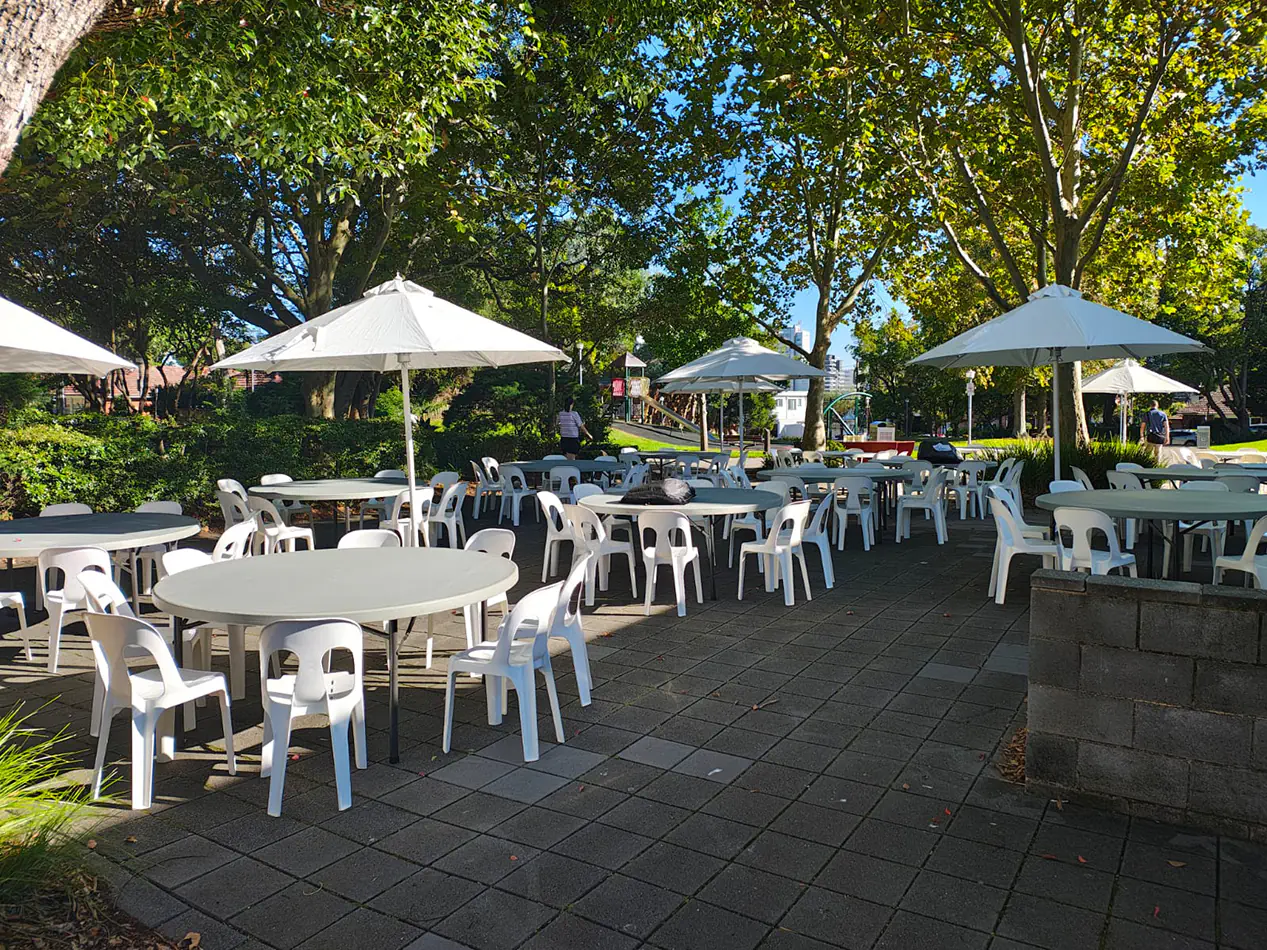 Outdoor event setup with round tables, white chairs and large umbrellas providing shade in a park environment with trees and paved area