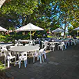 Outdoor event setup with round tables, white chairs and large umbrellas providing shade in a park environment with trees and paved area