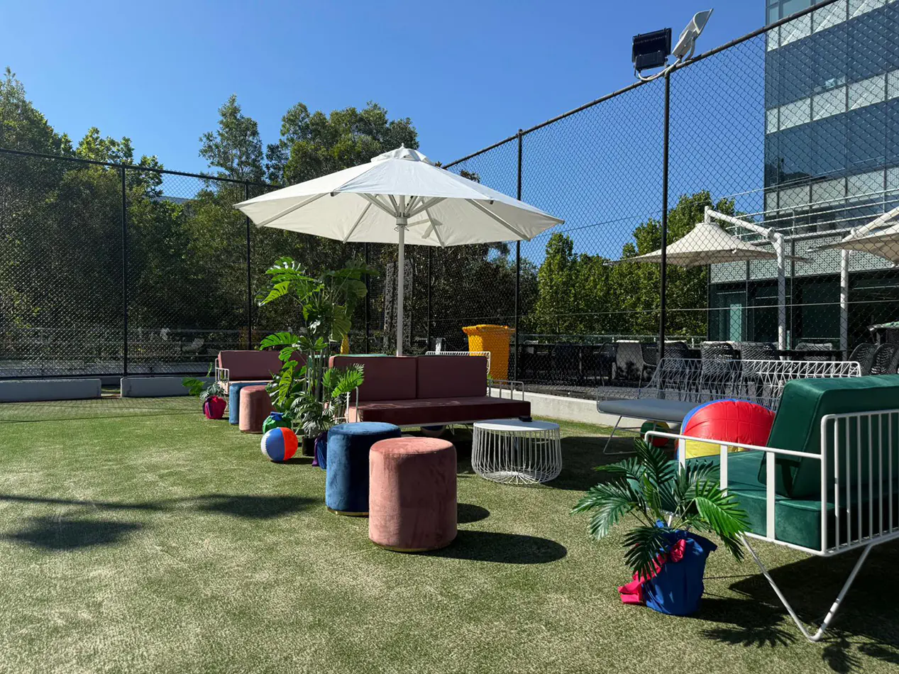 Outdoor lounge setting with velvet ottomans, upholstered sofas, coffee table and white umbrella on artificial grass with decorative plants