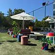 Outdoor lounge setting with velvet ottomans, upholstered sofas, coffee table and white umbrella on artificial grass with decorative plants