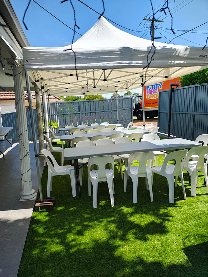 Outdoor marquee setup with long trestle tables and white plastic chairs arranged on artificial grass in a backyard setting