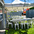 Outdoor marquee setup with long trestle tables and white plastic chairs arranged on artificial grass in a backyard setting