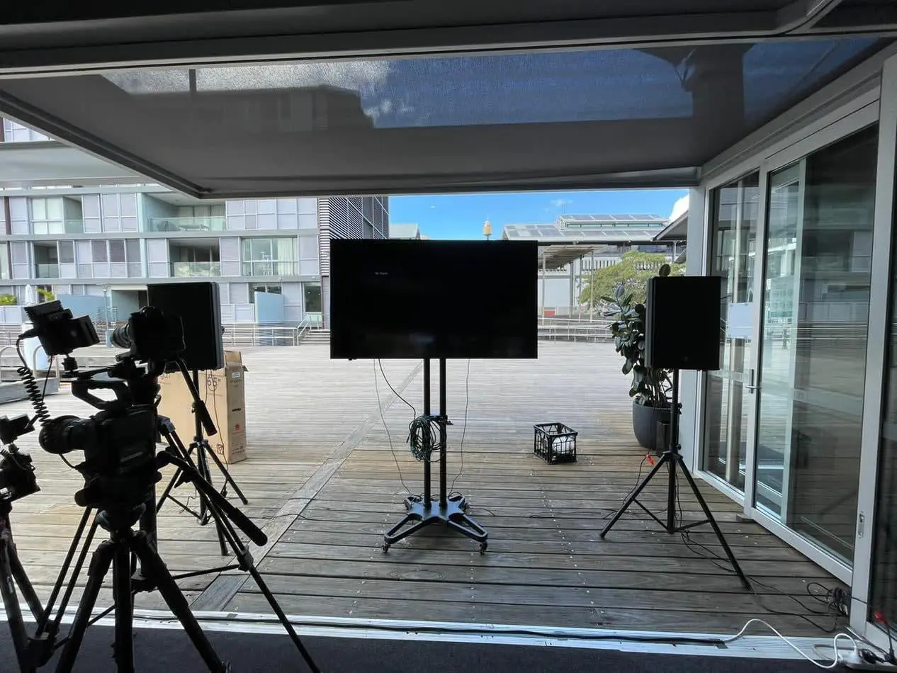 Outdoor event setup with TV screen on stand, speakers on tripods, and camera equipment arranged on timber deck balcony