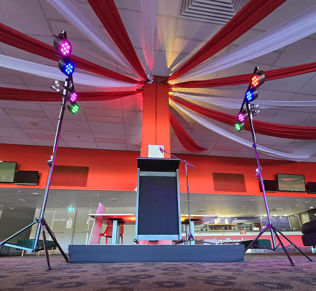 Event stage setup with podium and microphone, featuring par can lighting on stands casting colourful red, blue, and green lights with draped ceiling fabric in a function venue