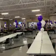 Description (modified): Purple and white balloons decorating plastic trestle tables in a school cafeteria.