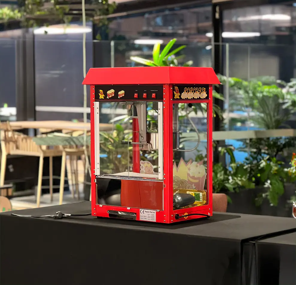 Close-up of a red commercial popcorn machine on a table in an indoor venue with plants and ambient lighting in the background