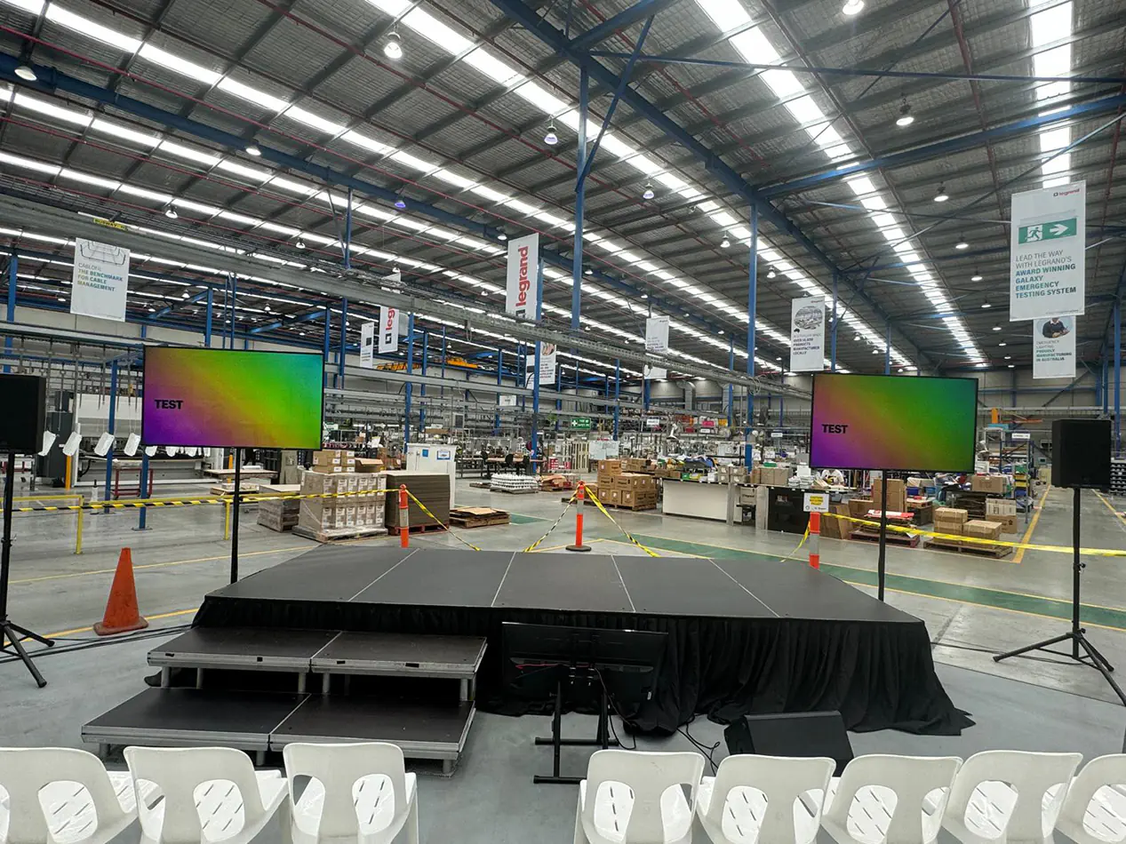 Portable event stage setup inside a large warehouse with dual presentation screens, speakers, stairs access, and rows of audience seating