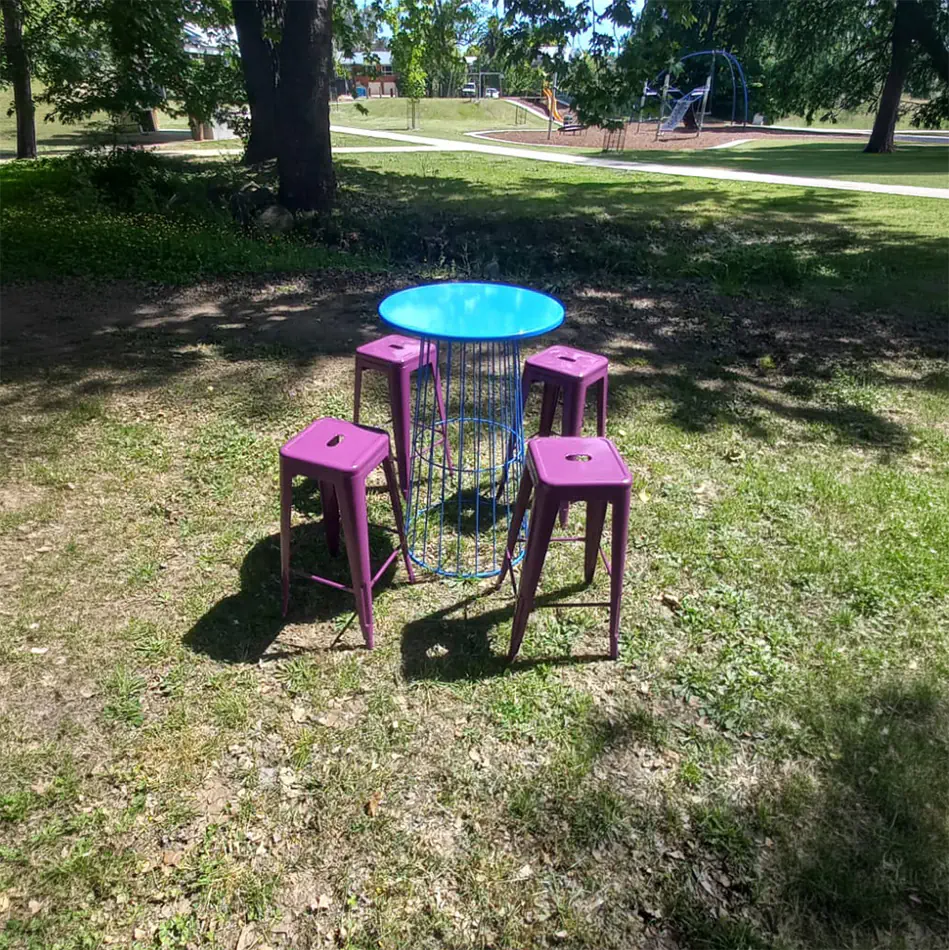 Purple Tolix stools arranged around a blue wire base cocktail table in an outdoor park setting with trees, grass, and playground in the background
