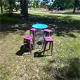Purple Tolix stools arranged around a blue wire base cocktail table in an outdoor park setting with trees, grass, and playground in the background