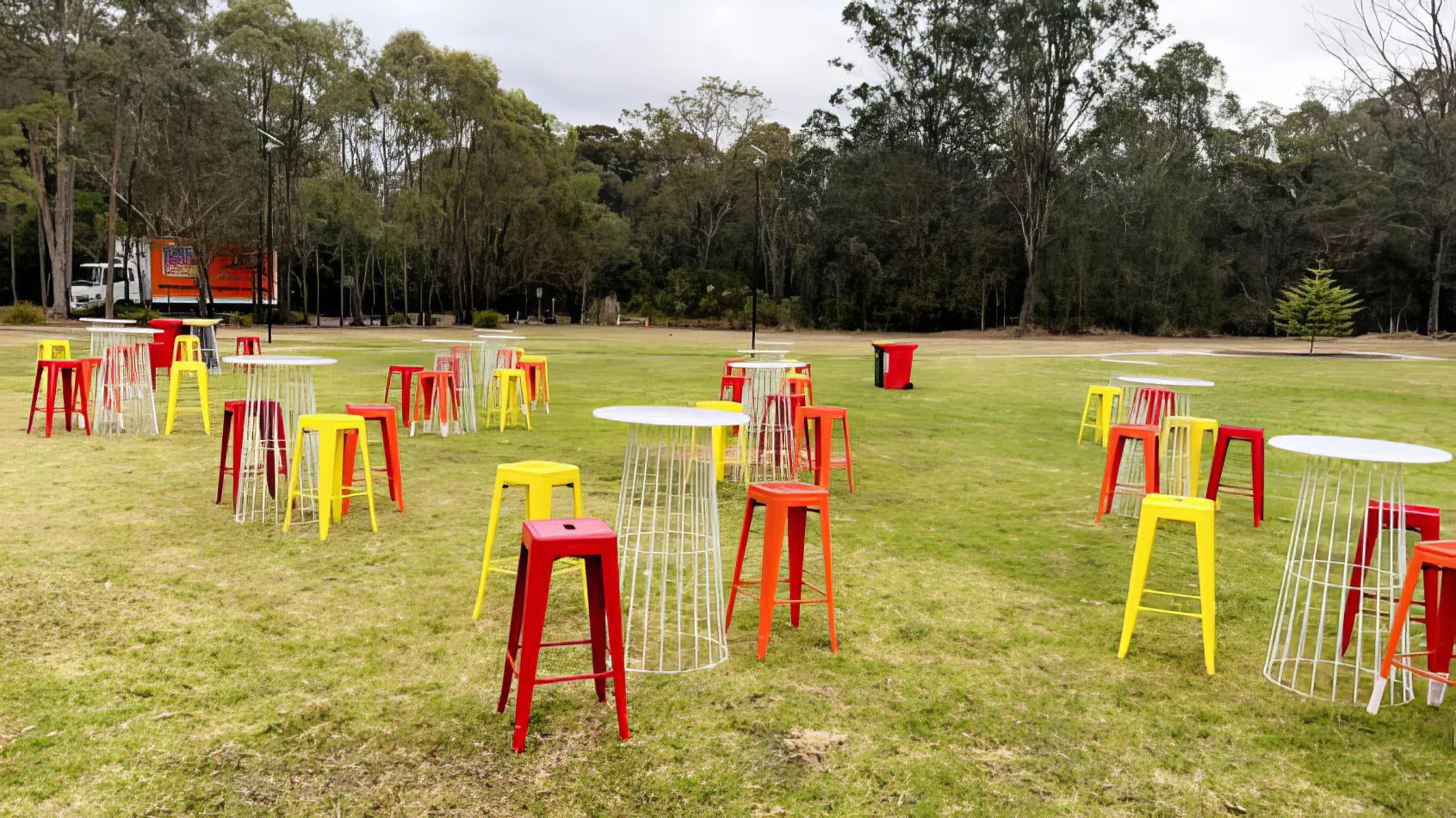 Red tolix stool hire mixed with yellow stools and wire cocktail tables arranged across a large outdoor grass event space with trees and open surroundings