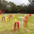 Red tolix stool hire mixed with yellow stools and wire cocktail tables arranged across a large outdoor grass event space with trees and open surroundings