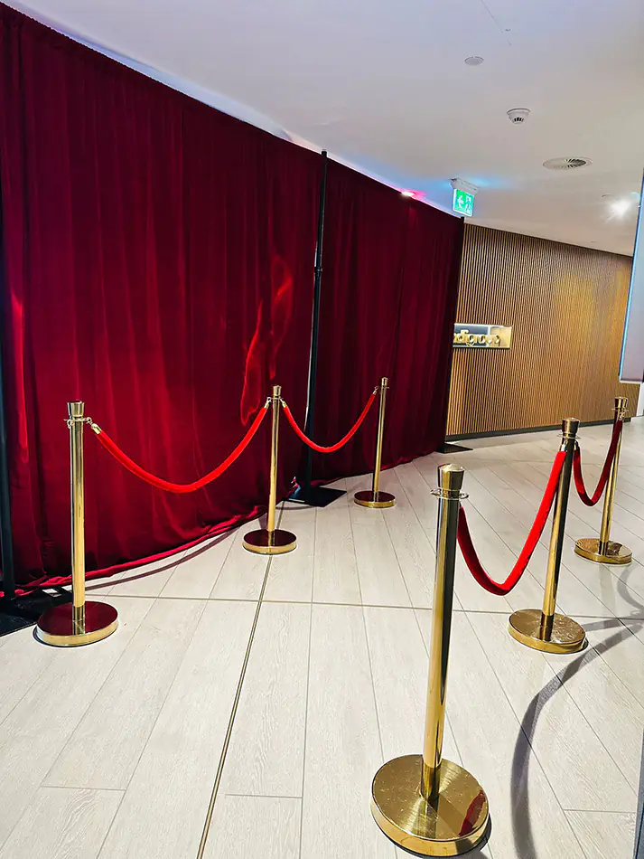 Red velvet draping with gold bollards and red ropes creating a formal event entry area inside a modern venue
