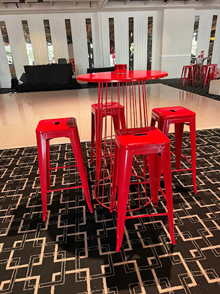 Red wire cocktail table with matching bar stools positioned in an indoor event space with patterned carpet flooring