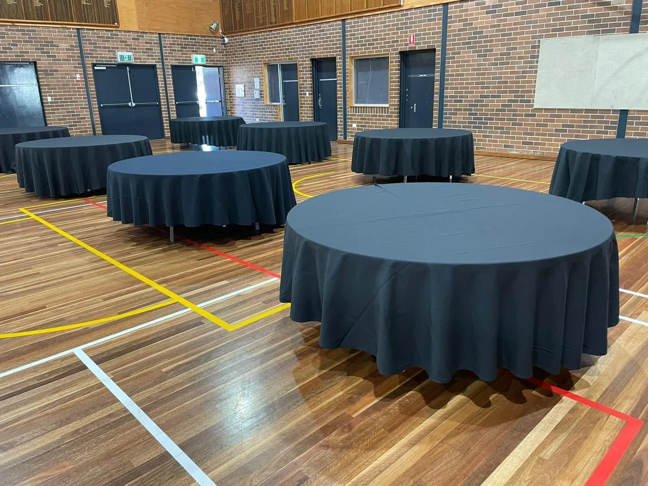 Tables and chairs, including round timber banquet tables, are set up in a gymnasium.