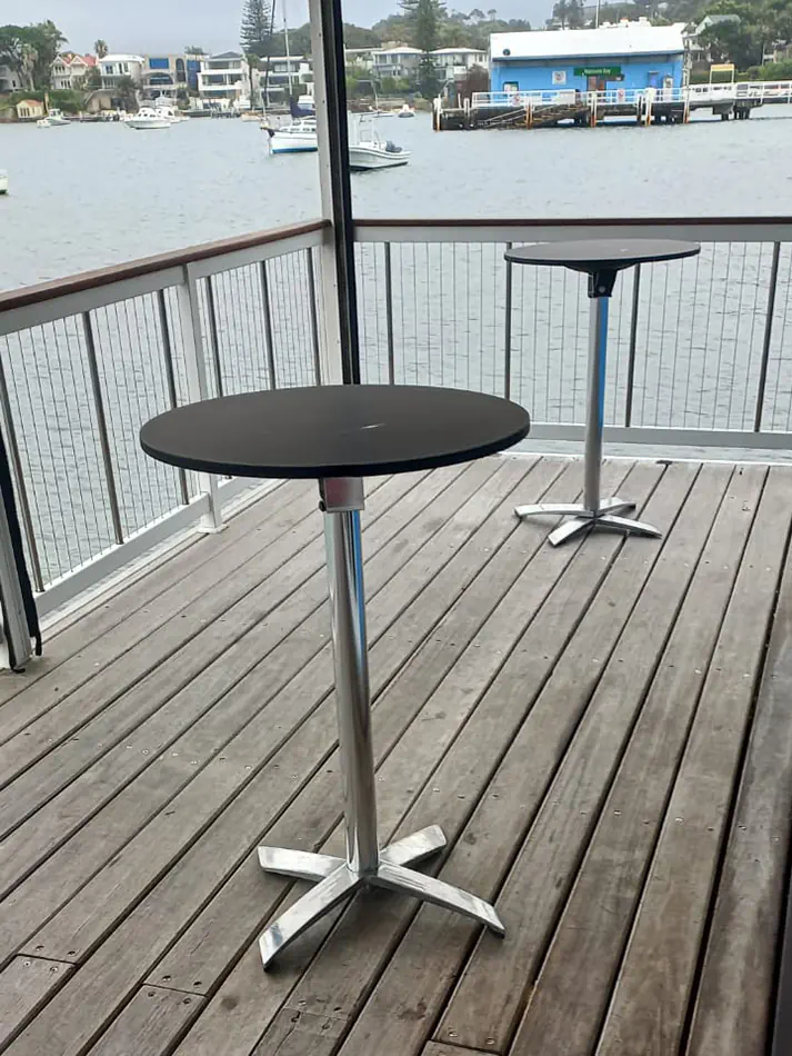 Round black cocktail tables with metal bases positioned on a timber deck overlooking a waterfront with boats and residential buildings in the background