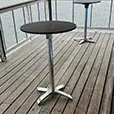 Round black cocktail tables with metal bases positioned on a timber deck overlooking a waterfront with boats and residential buildings in the background