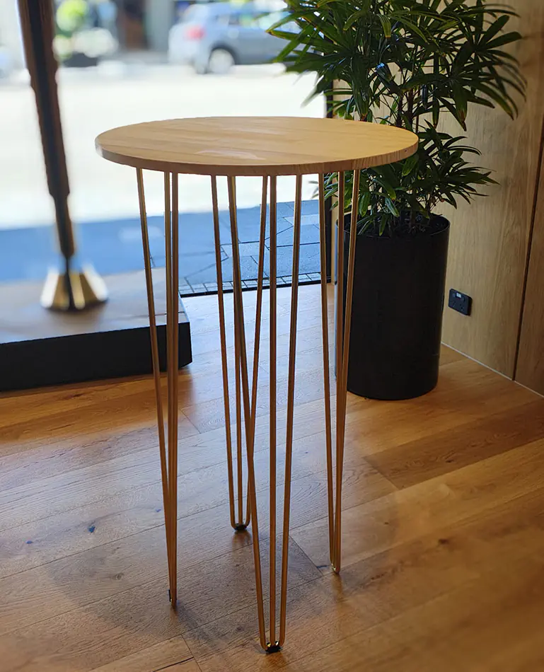 Round wooden cocktail table with gold hairpin legs positioned indoors on timber flooring beside a potted plant and window