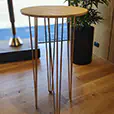 Round wooden cocktail table with gold hairpin legs positioned indoors on timber flooring beside a potted plant and window
