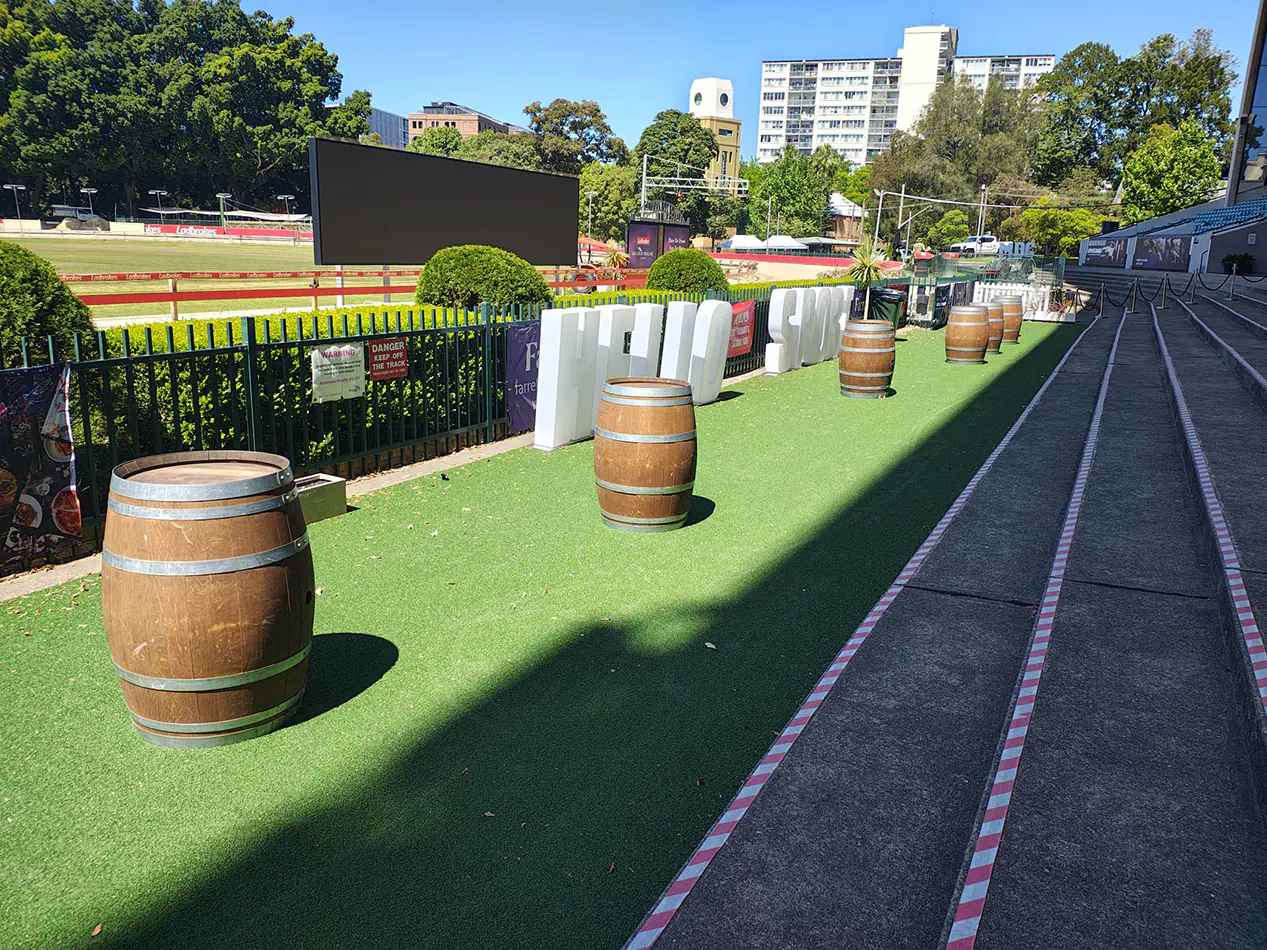 Rustic timber wine barrels arranged along a racecourse event walkway with artificial turf and outdoor entertainment area