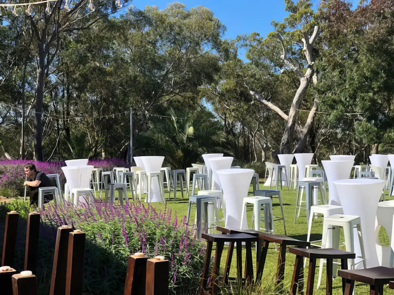 Silver tolix stool hire arranged with white cocktail tables across an outdoor garden event setting, surrounded by greenery, trees and decorative lighting
