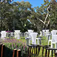 Silver tolix stool hire arranged with white cocktail tables across an outdoor garden event setting, surrounded by greenery, trees and decorative lighting