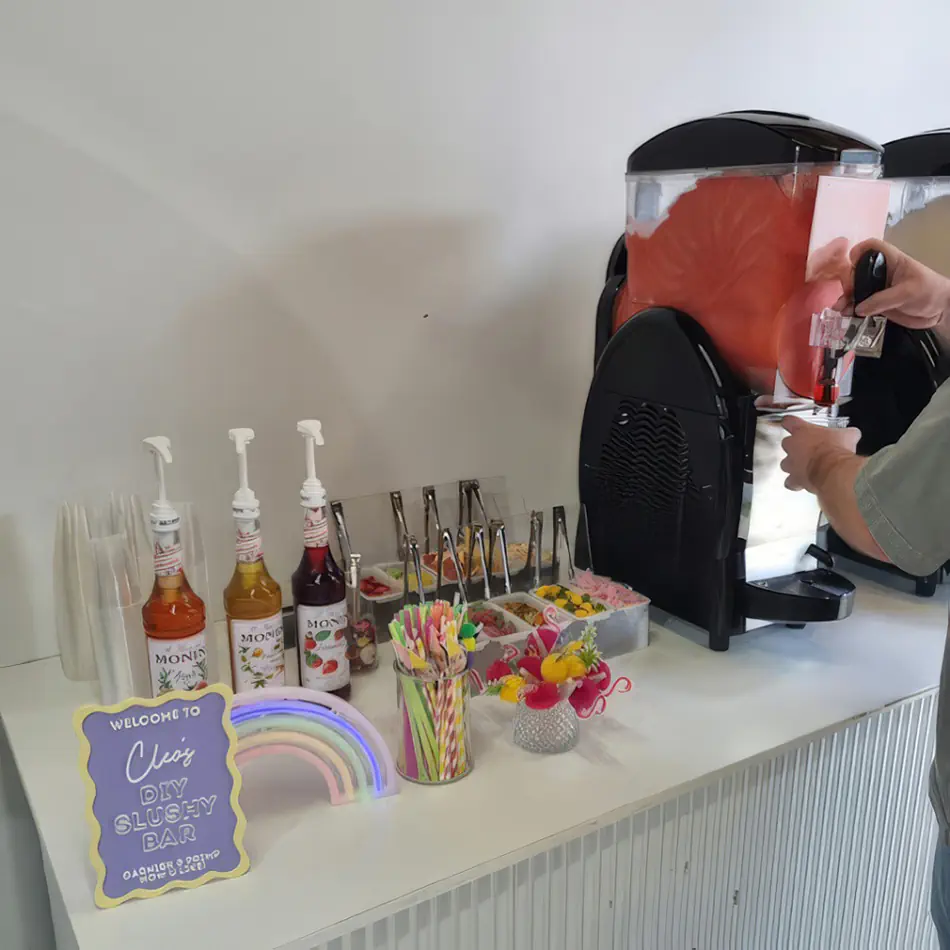 Slushie machine hire setup at a party drink station with flavoured syrups, cups, straws and a frozen drink dispenser in use