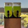 Two green juice dispensers on a table in front of a window. Slushie Machine Hire: Pkg 3 - *240 Drinks*