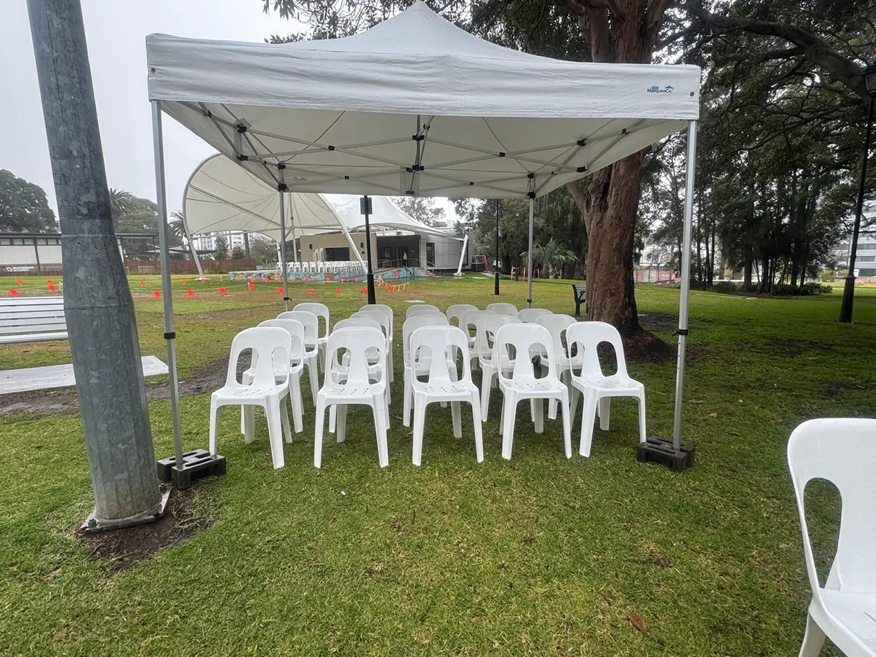 Small white pop-up marquee with rows of white event chairs set up in a public park for an outdoor gathering