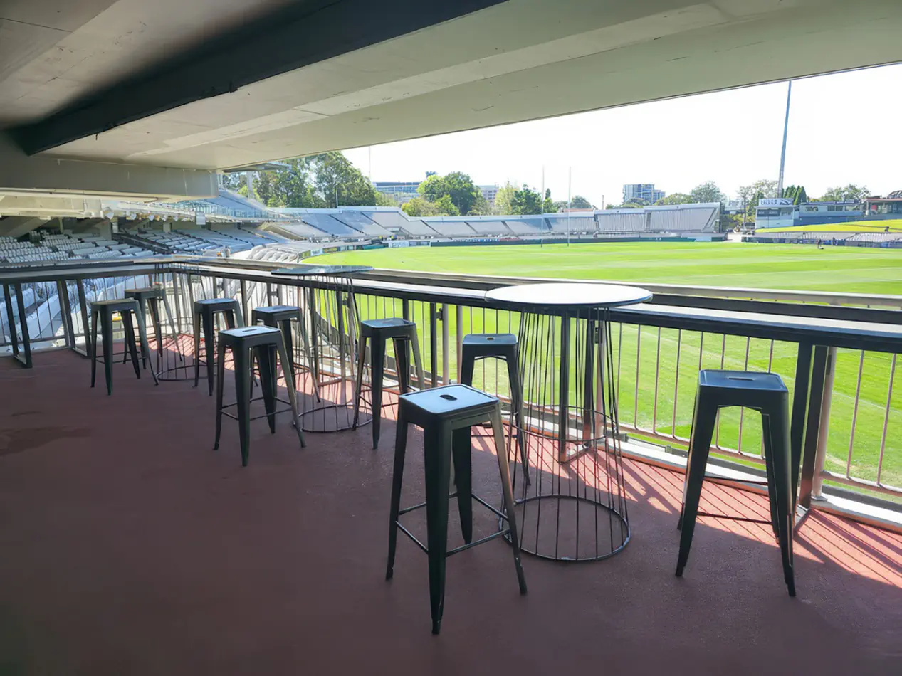 Stadium balcony seating area with black metal tolix stools and round tables overlooking a sports field in a covered viewing area