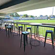 Stadium balcony seating area with black metal tolix stools and round tables overlooking a sports field in a covered viewing area