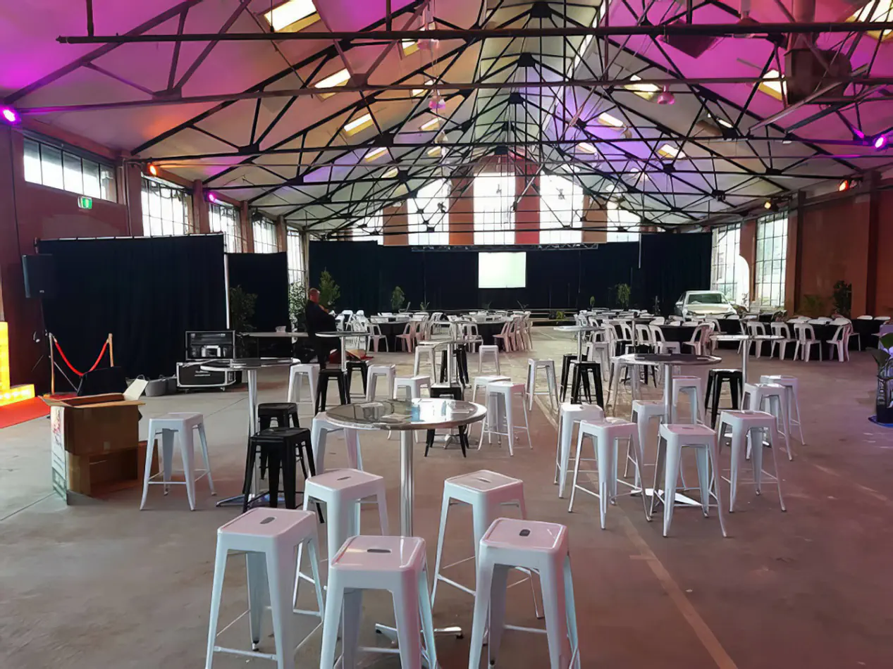 Multiple stainless steel cocktail tables arranged throughout a large function hall with white stools, stage area, and event lighting setup