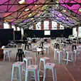 Multiple stainless steel cocktail tables arranged throughout a large function hall with white stools, stage area, and event lighting setup