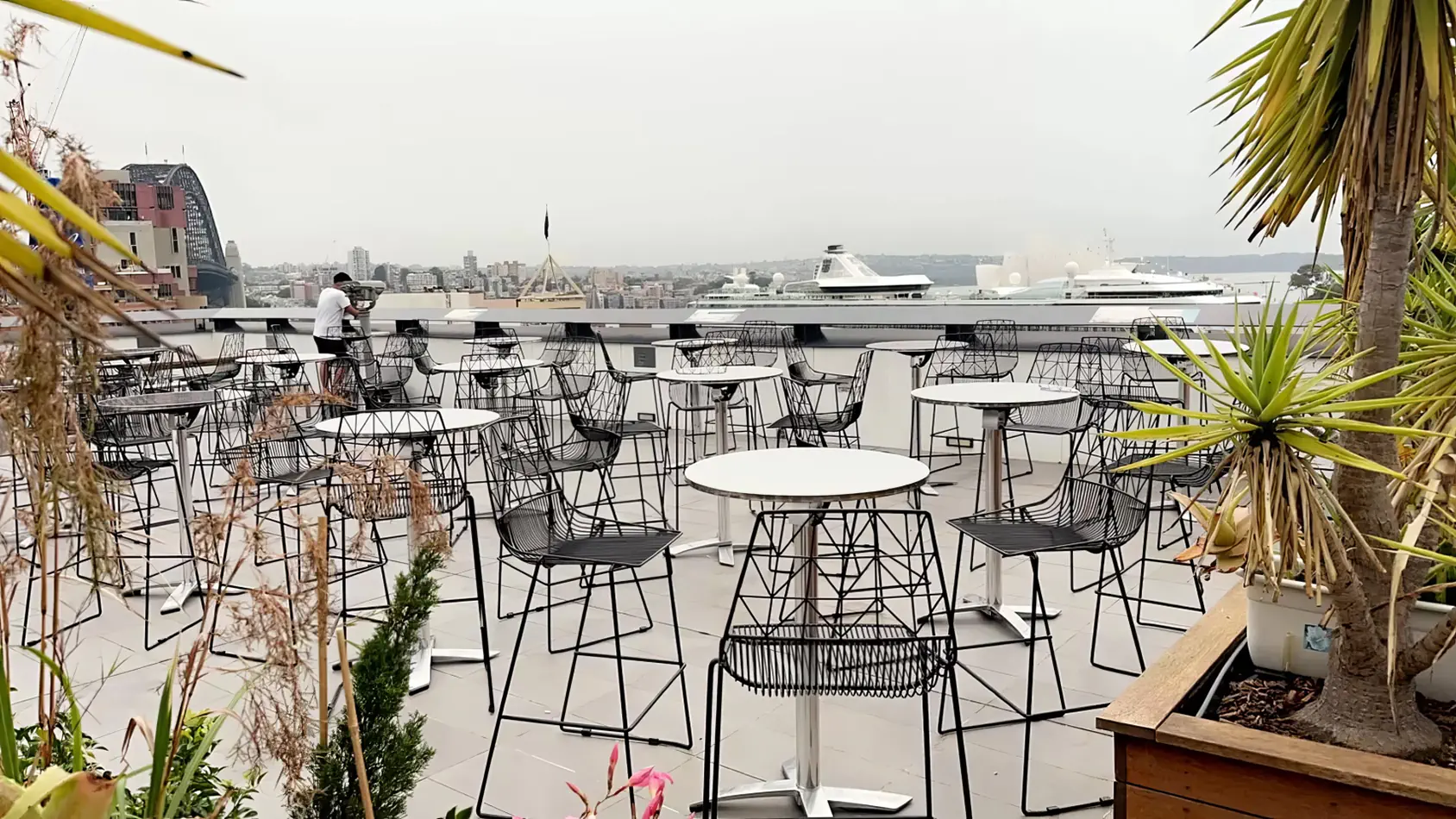 Stainless steel cocktail tables set across a rooftop terrace with black wire chairs and city skyline views in the background