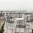 Stainless steel cocktail tables set across a rooftop terrace with black wire chairs and city skyline views in the background