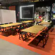 Natural timber-top banquet tables with black hairpin legs arranged in rows with matching black chairs, set up on an orange floor covering inside a retail venue for an organised event or group activity.