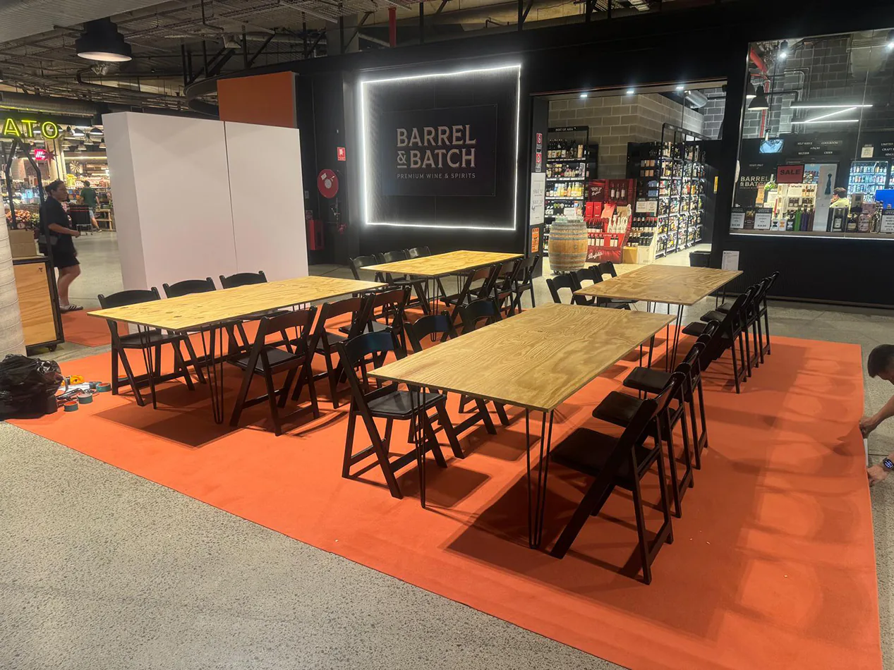 Natural timber-top banquet tables with black hairpin legs arranged in rows with matching black chairs, set up on an orange floor covering inside a retail venue for an organised event or group activity.