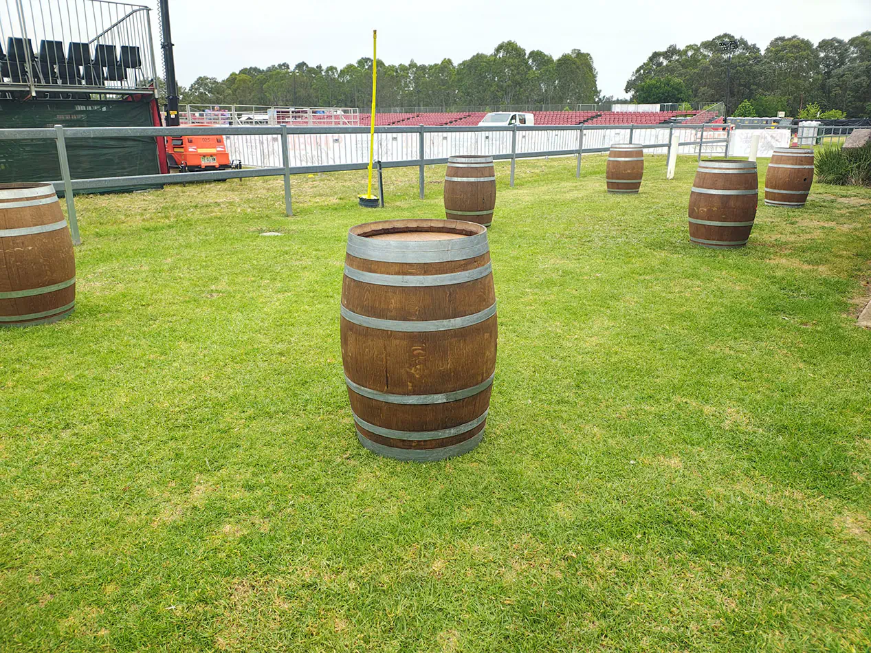 Timber wine barrels spaced across an outdoor event lawn beside stadium seating and fenced entertainment area