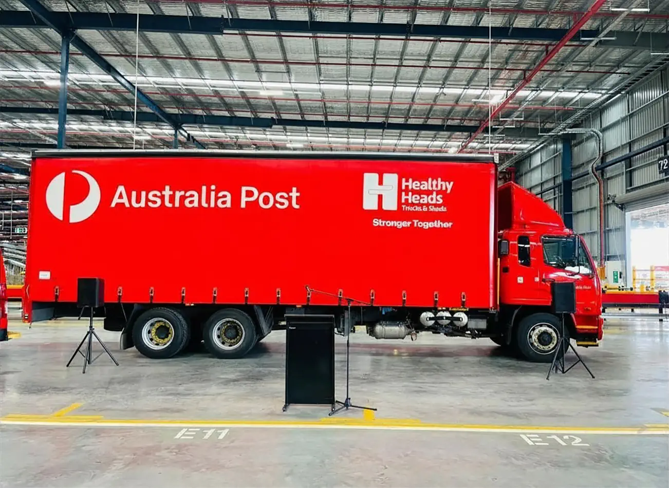 TV hire setup for an Australia Post corporate event inside a warehouse venue, with a freestanding display screen, microphone stand and PA speakers positioned near a branded truck.