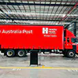 TV hire setup for an Australia Post corporate event inside a warehouse venue, with a freestanding display screen, microphone stand and PA speakers positioned near a branded truck.