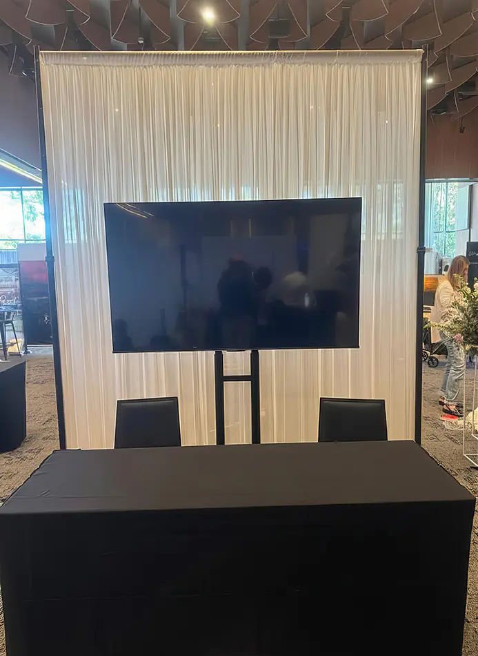 TV hire display screen positioned in front of a white draped backdrop with table and seating, set up for an indoor event or presentation area