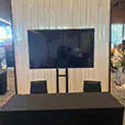 TV hire display screen positioned in front of a white draped backdrop with table and seating, set up for an indoor event or presentation area