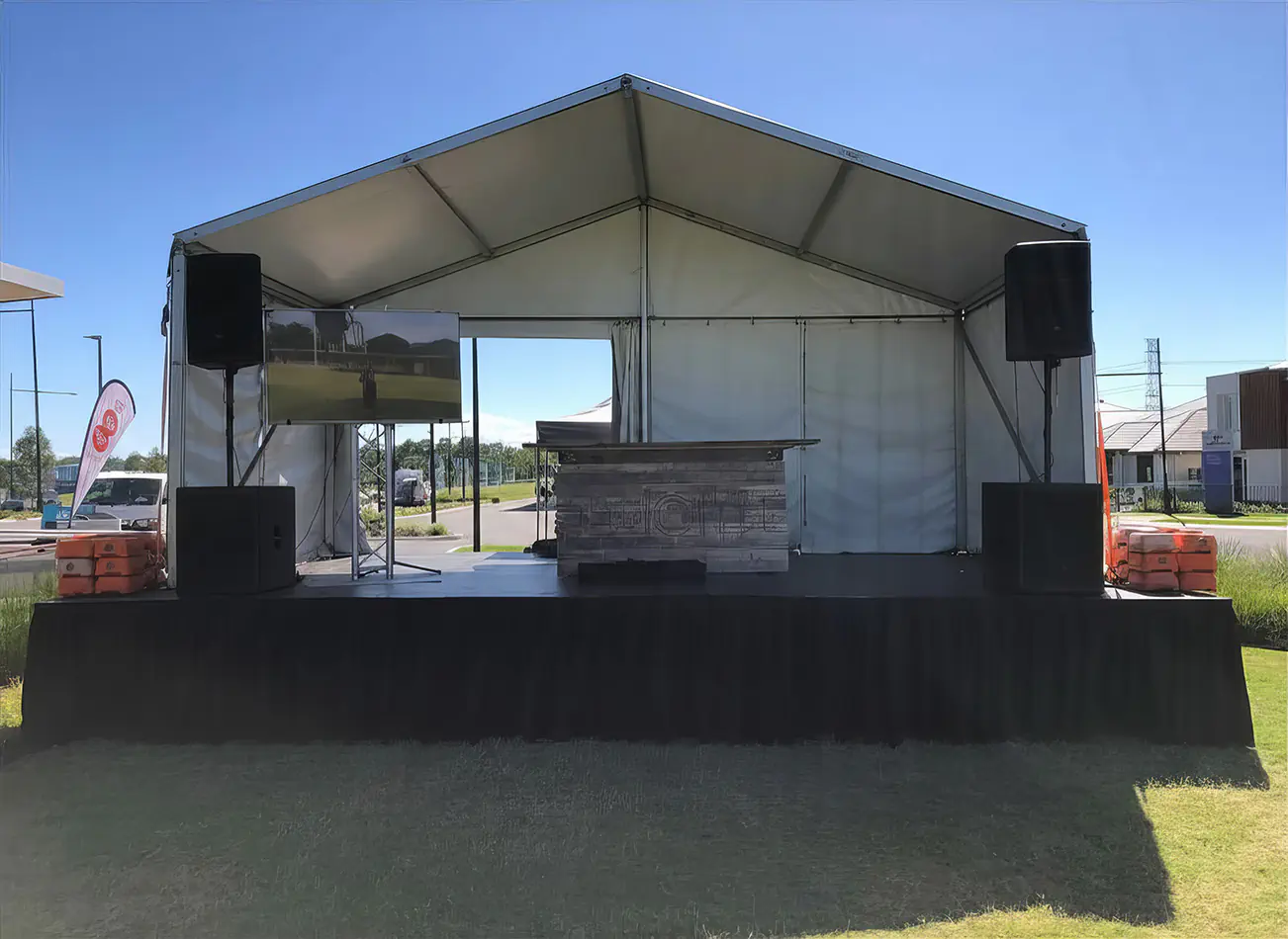 TV hire setup on an outdoor event stage under a marquee with PA speakers, display screen on stand and bar counter, set up for a corporate or community event