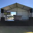 TV hire setup on an outdoor event stage under a marquee with PA speakers, display screen on stand and bar counter, set up for a corporate or community event