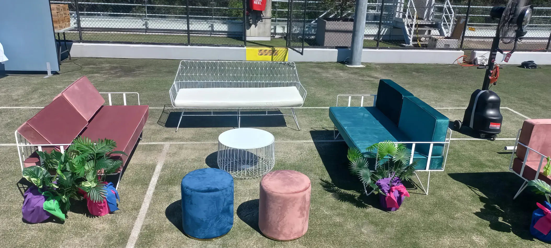 Outdoor lounge seating setup with green and pink velvet sofas, matching ottomans and a round coffee table arranged on artificial grass