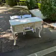 Elegant vintage table with carved drawer accents and cabriole legs, set alongside two white chairs in a landscaped outdoor garden.