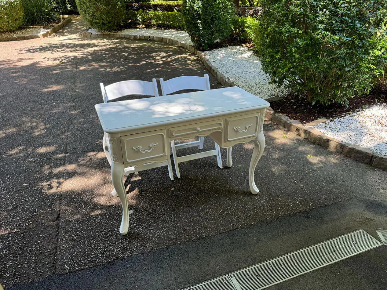 Elegant vintage table with carved drawer accents and cabriole legs, set alongside two white chairs in a landscaped outdoor garden.